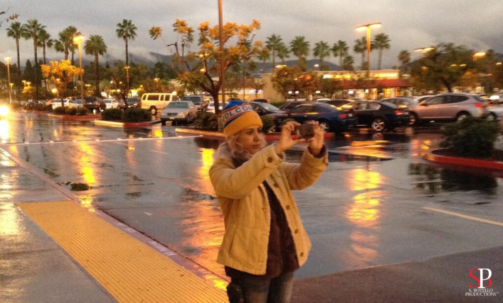 Susy Botello capturing a sunset in the rain with palm trees wearing ski hat and jacket, holding a smartphone horizontally.